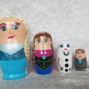 May include: Set of five hand-painted wooden nesting dolls featuring characters from the animated film Frozen. The largest doll is a blonde woman with a blue dress and a sparkly bodice. The next doll is a brunette woman with a pink and blue dress. The third doll is a white snowman with a black top hat and black buttons. The fourth doll is a man with brown hair and a gray jacket. The smallest doll is a brown bird.