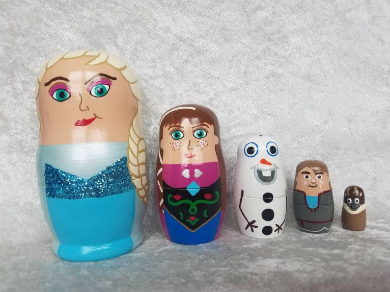 May include: Set of five hand-painted wooden nesting dolls featuring characters from the animated film Frozen. The largest doll is a blonde woman with a blue dress and a sparkly bodice. The next doll is a brunette woman with a pink and blue dress. The third doll is a white snowman with a black top hat and black buttons. The fourth doll is a man with brown hair and a gray jacket. The smallest doll is a brown bird.