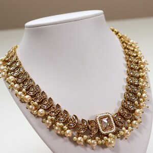 May include: A gold necklace featuring a large, clear, rectangular stone at its centre. The necklace is adorned with smaller stones and pearls.