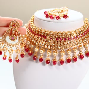 May include: A gold necklace and earrings set with red and white stones. The necklace has a thick, layered design with dangling pearls and red beads. The earrings are teardrop shaped with a similar design.