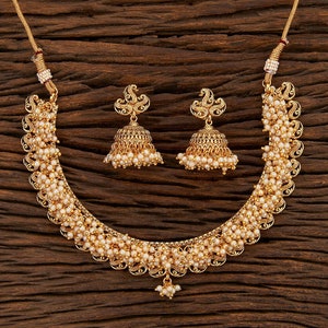 Op de afbeelding: Een goudkleurige ketting- en oorbellenset met kleine, ronde, cr&egrave;mekleurige kralen. De ketting heeft een gebogen ontwerp met ingewikkelde details en de oorbellen zijn klokvormig met vergelijkbare kraalaccenten. De sieradenset wordt gepresenteerd op een houten oppervlak.