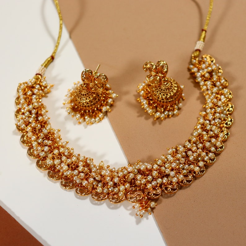 Op de afbeelding: Een goudkleurige ketting en bijpassende oorbellen set. De ketting en oorbellen zijn versierd met kleine, ronde, witte parels. De ketting heeft een geschulpte vormgeving met ingewikkelde details. De oorbellen zijn rond met een vergelijkbaar parel- en goudontwerp.