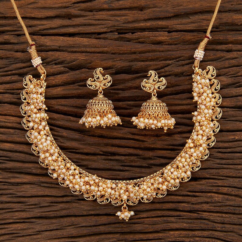 Op de afbeelding: Een goudkleurige ketting- en oorbellenset met kleine, ronde, cr&egrave;mekleurige kralen. De ketting heeft een gebogen ontwerp met ingewikkelde details en de oorbellen zijn klokvormig met vergelijkbare kraalaccenten. De sieradenset wordt gepresenteerd op een houten oppervlak.