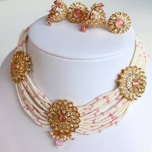 May include: A white pearl and pink bead necklace with gold accents and pink stones. The necklace has multiple strands and features a large floral pendant with a pink stone in the centre. Matching earrings are also included.