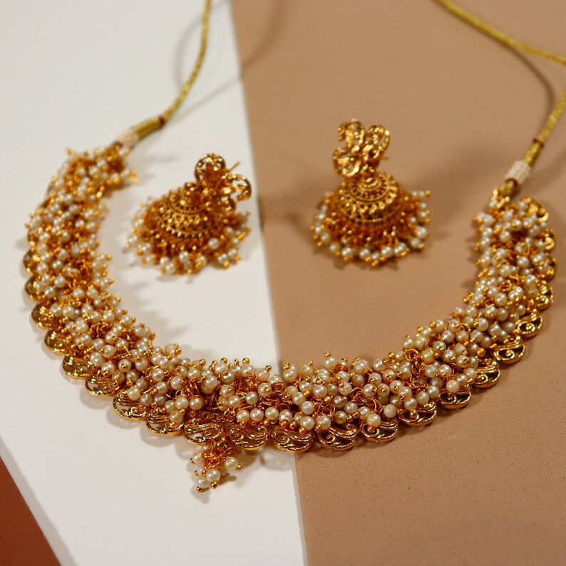 Op de afbeelding: Een gouden ketting en bijpassende oorbellen set. De ketting heeft gouden filigraan elementen en kleine, witte parels. De oorbellen hebben een vergelijkbaar ontwerp, met gouden details en parels.