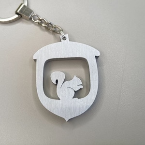 Puede incluir: Un llavero de plata con una silueta de ardilla dentro de una forma de bellota.