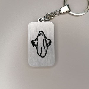 Op de afbeelding: Een rechthoekige zilveren sleutelhanger met een zwart spookontwerp. Het spook is een eenvoudige omtrek met twee ovale ogen en een vloeiend lichaam. De sleutelhanger heeft een zilveren ketting en een ronde sleutelring.