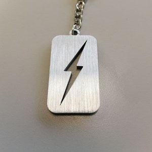 Può includere: Portachiavi in ​​argento con un design a forma di fulmine tagliato. Il portachiavi è attaccato a un anello e una catena d'argento.