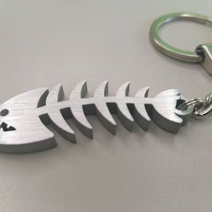 Puede incluir: Un llavero de hueso de pescado plateado con una cadena plateada y un anillo plateado. El hueso de pescado está detallado con una boca y costillas.