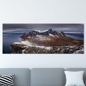 May include: A panoramic landscape print of a mountain range with snow-capped peaks, overlooking a dark blue body of water. The artwork is displayed above a gray sectional sofa with decorative pillows, set against a white wall and light wood floor.