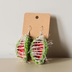Puede incluir: Un par de pendientes de color verde y rojo inspirados en la sandía con una textura tejida. Los pendientes cuelgan de una tarjeta marrón.