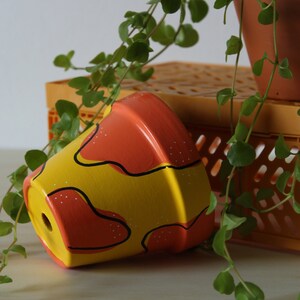 May include: A hand-painted ceramic flower pot with a yellow base and orange top. The pot features a wavy design in orange and black, with small white dots. Green vines and a terracotta pot are in the background.
