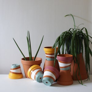 Op de afbeelding: Een verzameling terracotta bloempotten, sommige rechtop en sommige gekanteld, met geschilderde horizontale strepen in verschillende kleuren, waaronder geel, roze en blauwgroen. Verschillende potten bevatten groene planten.