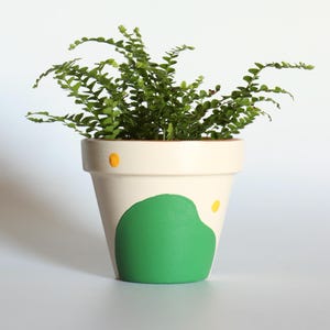 May include: A small potted fern with bright green foliage. The off-white ceramic pot is decorated with a large green shape and two yellow dots. The plant is set against a plain white background.