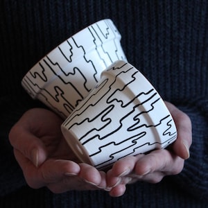Puede incluir: Una taza de cerámica blanca con un asa negra y el texto "But First, Coffee" en negro, en negrita. La taza está sobre una superficie blanca.