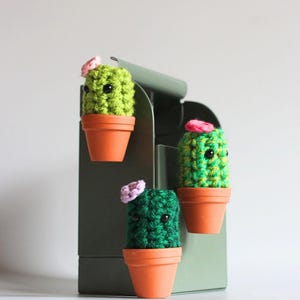 May include: Three crocheted cactus figures in small terracotta-coloured pots attached to a sage green metal mailbox. Each cactus is a different shade of green, features two black bead eyes, and is topped with a small pink or purple flower.