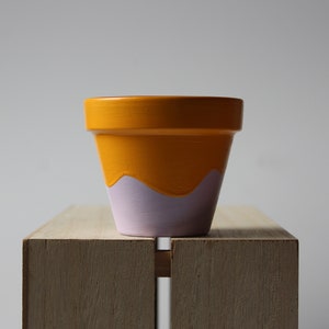 Peut inclure: Un petit pot en terre cuite peint avec un motif jaune et violet. Le pot est posé sur une surface en bois.