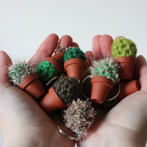Pode incluir: Uma coleção de chaveiros de cacto de crochê em vários tons de verde e marrom. Os cactos estão em vasos de terracota pequenos e têm anéis de metal para prender às chaves.