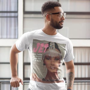 May include: White t-shirt featuring a vintage magazine cover with the word "JET" in pink and a portrait of a woman wearing a jeweled headpiece. The text "Ebony Fashion Fair Presents Hottest Show in 26 Years" is also visible.