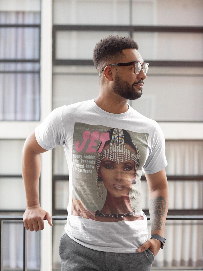May include: White t-shirt featuring a vintage magazine cover with the word "JET" in pink and a portrait of a woman wearing a jeweled headpiece. The text "Ebony Fashion Fair Presents Hottest Show in 26 Years" is also visible.