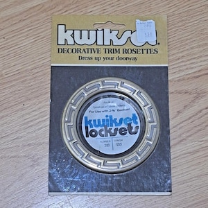 May include: A Kwikset decorative trim rosette in a gold-tone finish with a Greek key pattern, packaged on a cardboard backing. The packaging features the text Kwikset Decorative Trim Rosettes, fits most cylindrical or tubular locksets, and 2-3/8 inch backset.