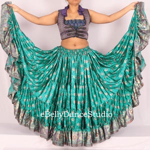 May include: A teal and gold belly dance skirt with a matching vest. The skirt has multiple layers of fabric with a floral pattern. The vest is purple and black with a decorative design. The eBellyDanceStudio logo is visible.