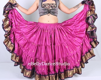 May include: A vibrant pink belly dance skirt with multiple ruffled tiers. The skirt has gold and navy blue trim along the edges. The top is a silver vest with a floral patterned top and a lace-up front. The text "eBellyDanceStudio" is visible.