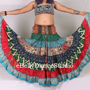 May include: A colourful, multi-layered skirt with a variety of floral and paisley patterns. The skirt is made of silk and has a flowing, bohemian style. The skirt is being held out by a person, showing off its full length and volume. The text "eBellyDanceStudio" is visible on the skirt.