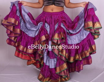 May include: A vibrant, layered belly dance skirt in shades of purple, lavender, and gold. The skirt features multiple tiers of fabric with gold trim and a flowing design. The text "eBellyDanceStudio" is visible.