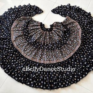 May include: Black belly dance skirt with a wide, flowing design. The skirt features a detailed pattern of small white flowers and a decorative waistband with a geometric design. The text "eBellyDanceStudio" is visible on the skirt.