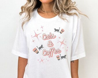 May include: White t-shirt with the text "Cats & Coffee" in a retro font, accompanied by illustrations of cats, coffee cups, and starbursts. The design is in shades of pink, brown, and black.