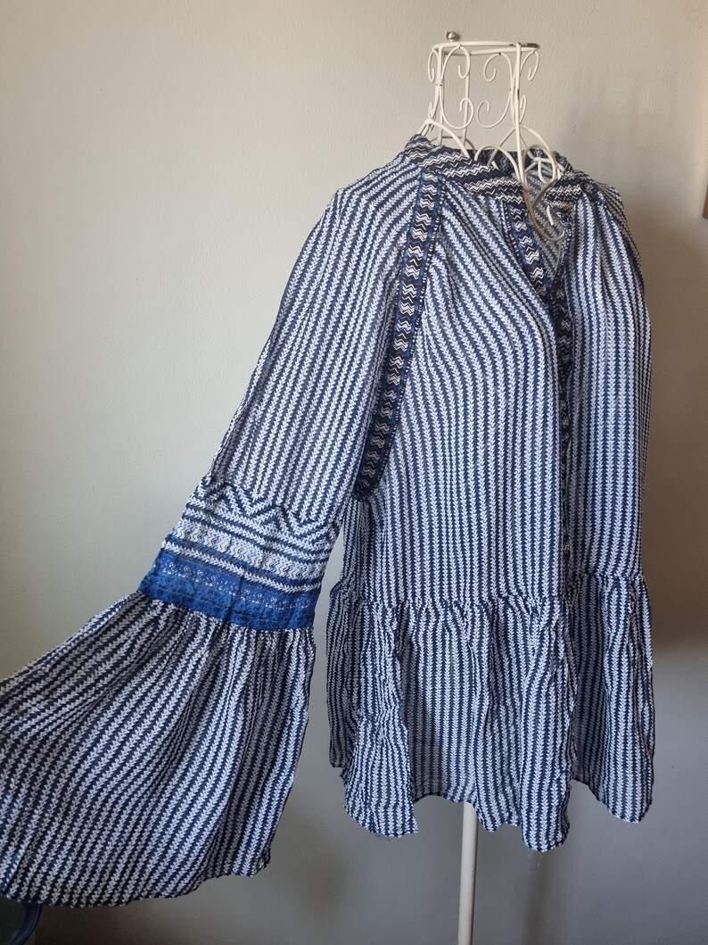May include: A long-sleeved blouse with a vertical striped pattern in navy blue and white. The sleeves have a flared design with blue lace trim. The blouse is displayed on a white mannequin.