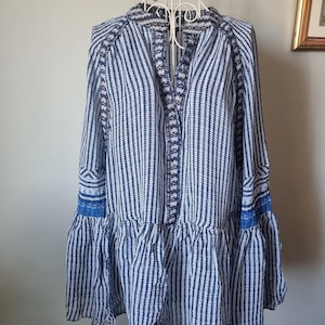 May include: A blue and white striped tunic-style top with long sleeves and a ruffled hem. The top features intricate embroidered details along the neckline and sleeve cuffs. The garment is displayed on a white dress form.