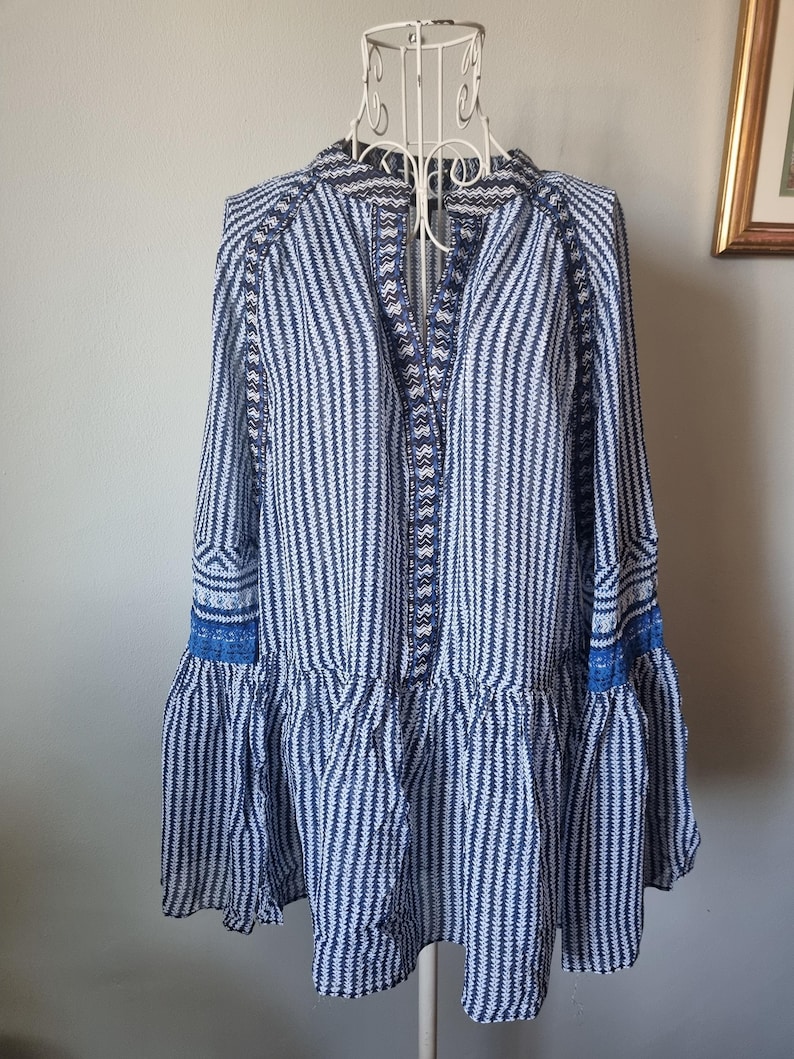 May include: A blue and white striped tunic-style top with long sleeves and a ruffled hem. The top features intricate embroidered details along the neckline and sleeve cuffs. The garment is displayed on a white dress form.