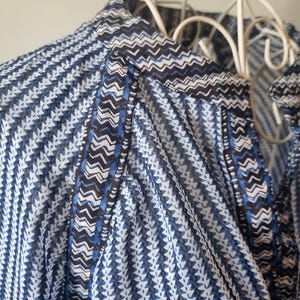 May include: Close-up of a blue and white patterned blouse. The fabric features a repeating geometric design with a mix of blue, white, and black accents. The blouse is hanging on a white metal hanger, showcasing the intricate pattern and lightweight material.