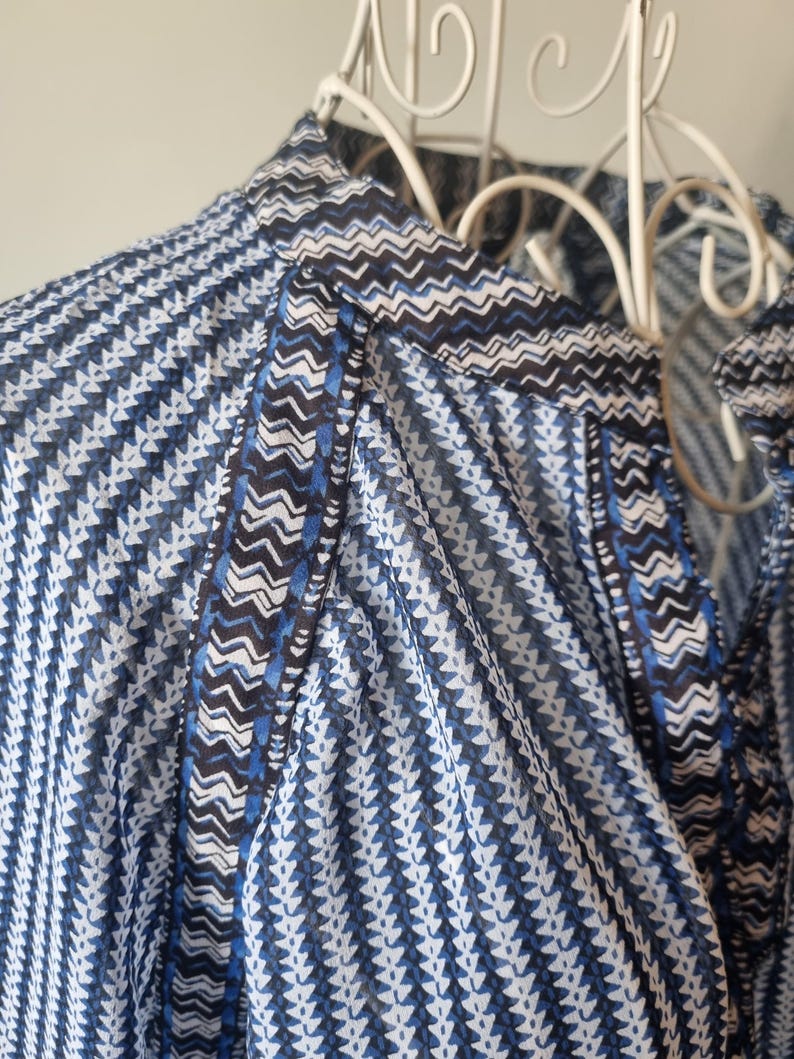 May include: Close-up of a blue and white patterned blouse. The fabric features a repeating geometric design with a mix of blue, white, and black accents. The blouse is hanging on a white metal hanger, showcasing the intricate pattern and lightweight material.