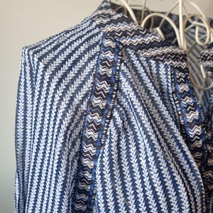 May include: Close-up of a blue and white patterned blouse. The fabric features a repeating chevron and vertical stripe design. The collar and trim have a contrasting dark blue and white zigzag pattern. The blouse is displayed on a white decorative hanger.