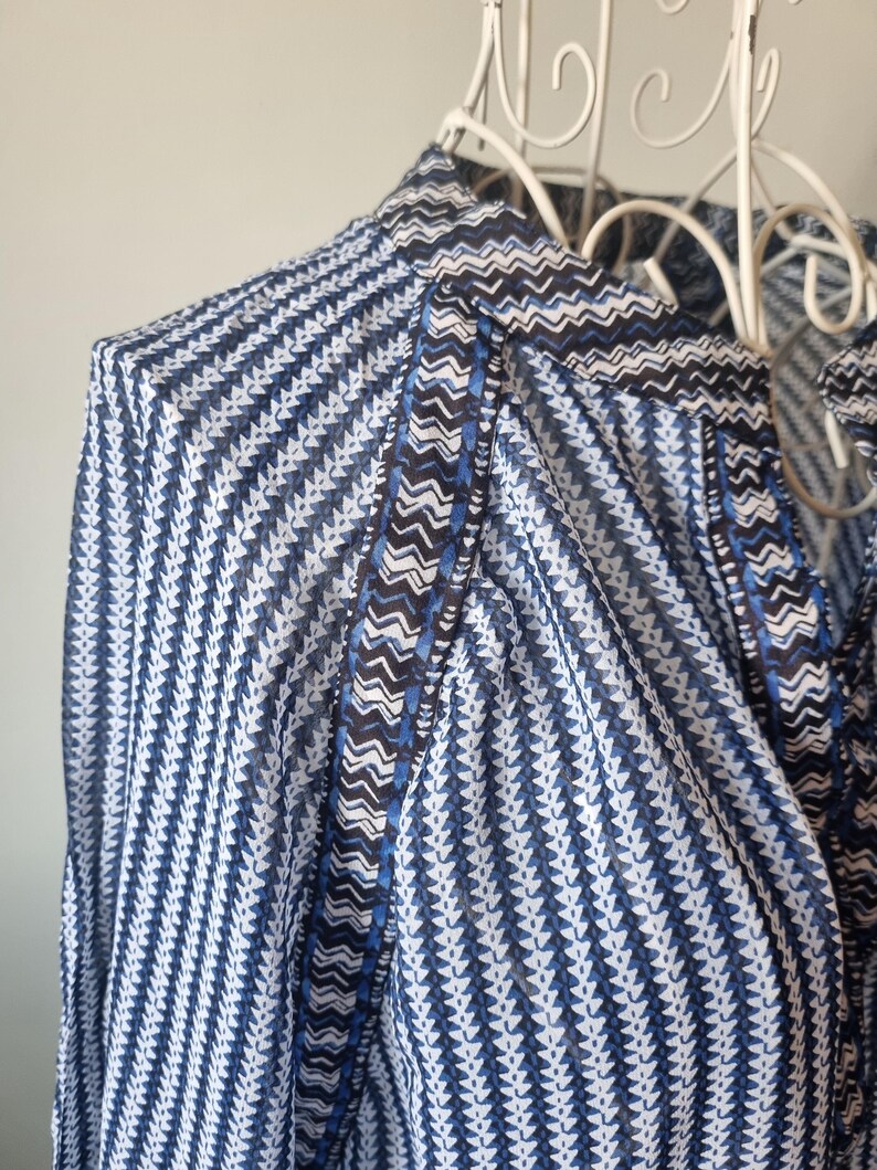 May include: Close-up of a blue and white patterned blouse. The fabric features a repeating chevron and vertical stripe design. The collar and trim have a contrasting dark blue and white zigzag pattern. The blouse is displayed on a white decorative hanger.