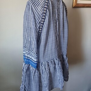 May include: A blue and white striped tunic dress with long sleeves and a ruffled hem. The dress features decorative blue trim on the sleeves and back. The dress is displayed on a white hanger.