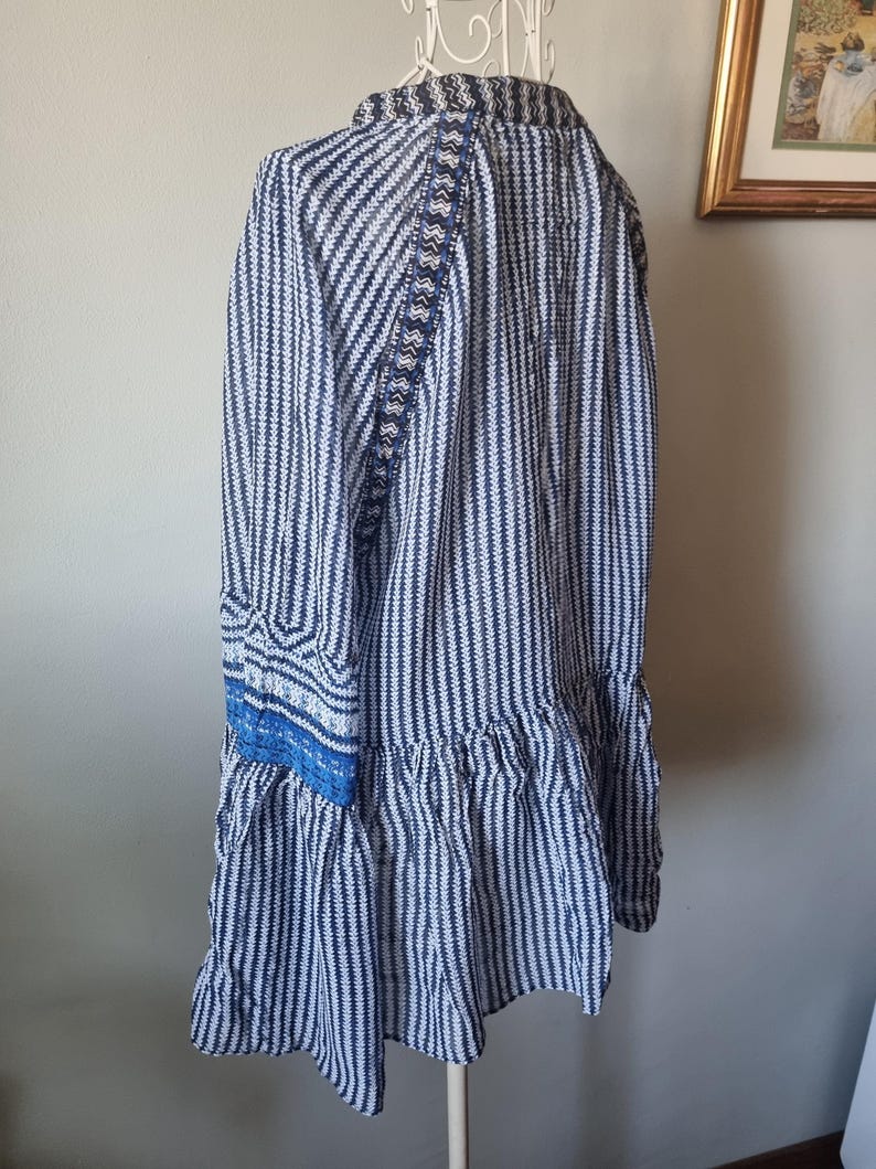 May include: A blue and white striped tunic dress with long sleeves and a ruffled hem. The dress features decorative blue trim on the sleeves and back. The dress is displayed on a white hanger.