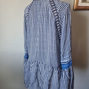 May include: A long-sleeved tunic with a vertical striped pattern in navy blue and white. The tunic features a ruffled hem and blue lace detailing on the sleeves. The garment is displayed on a white mannequin.