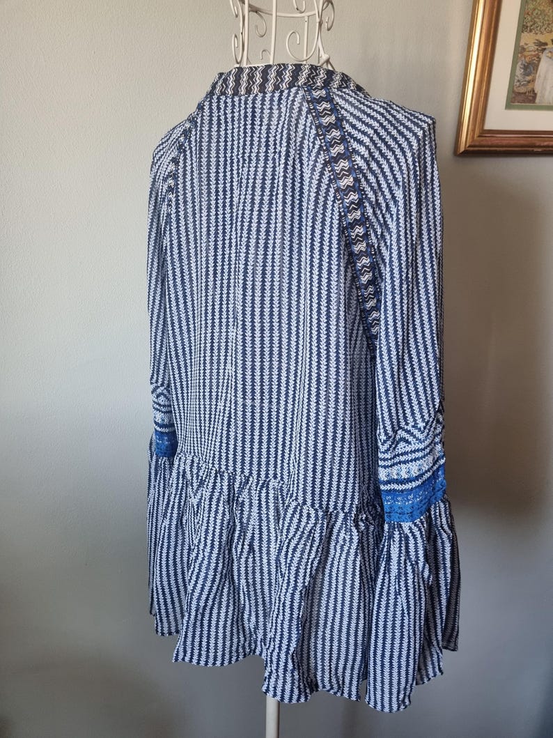 May include: A long-sleeved tunic with a vertical striped pattern in navy blue and white. The tunic features a ruffled hem and blue lace detailing on the sleeves. The garment is displayed on a white mannequin.