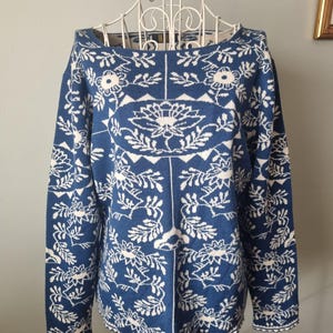 Pode incluir: Um suéter azul com um padrão floral branco. O suéter tem decote barco e mangas compridas. O design floral inclui flores e folhas. É exibido em um manequim branco.