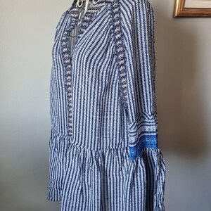 May include: A blue and white striped dress with long sleeves and a ruffled hem. The dress features a high collar and decorative blue trim along the neckline and cuffs. The dress is displayed on a white hanger.
