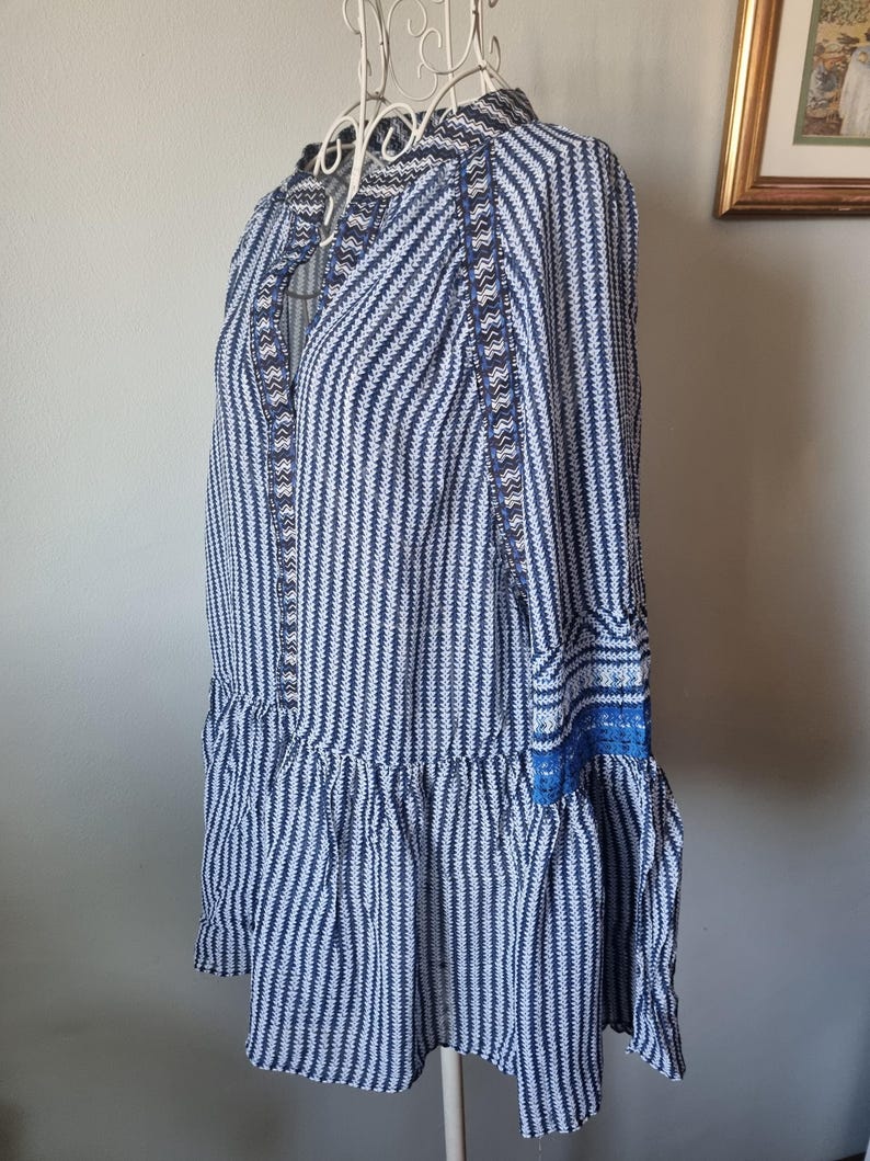 May include: A blue and white striped dress with long sleeves and a ruffled hem. The dress features a high collar and decorative blue trim along the neckline and cuffs. The dress is displayed on a white hanger.