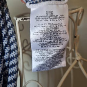 May include: A white tag with black text, attached to a garment with a blue and white floral pattern. The tag includes the RN and CA numbers, along with washing instructions and fabric composition details. The garment appears to be made of silk.