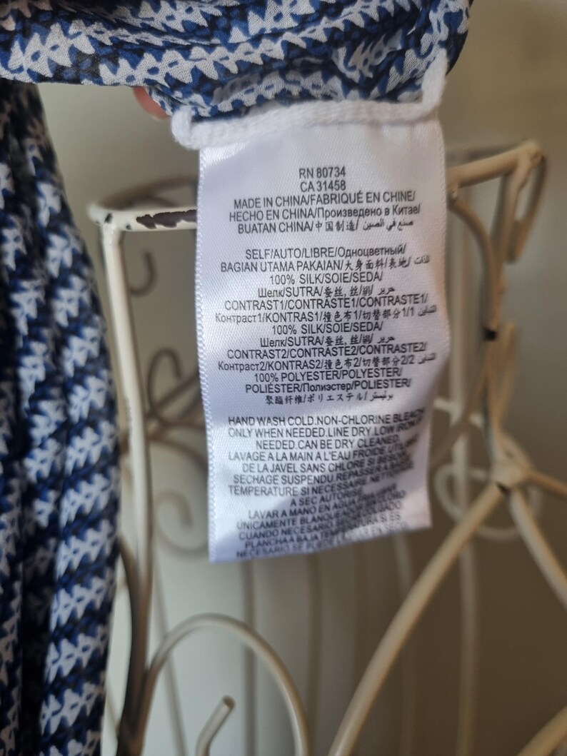 May include: A white tag with black text, attached to a garment with a blue and white floral pattern. The tag includes the RN and CA numbers, along with washing instructions and fabric composition details. The garment appears to be made of silk.