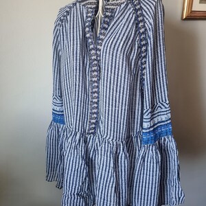 May include: A blue and white striped tunic dress with long sleeves and a ruffled hem. The dress features intricate detailing along the neckline and cuffs, with a mix of solid and patterned fabrics. The dress is displayed on a white metal hanger.