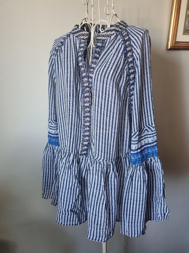 May include: A blue and white striped tunic dress with long sleeves and a ruffled hem. The dress features intricate detailing along the neckline and cuffs, with a mix of solid and patterned fabrics. The dress is displayed on a white metal hanger.