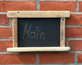 Può includere: Una piccola lavagna con la parola "Moin" e un simbolo a forma di cuore scritti con gesso bianco, incorniciata da un bordo in legno chiaro. La lavagna è montata su un muro di mattoni rossi.